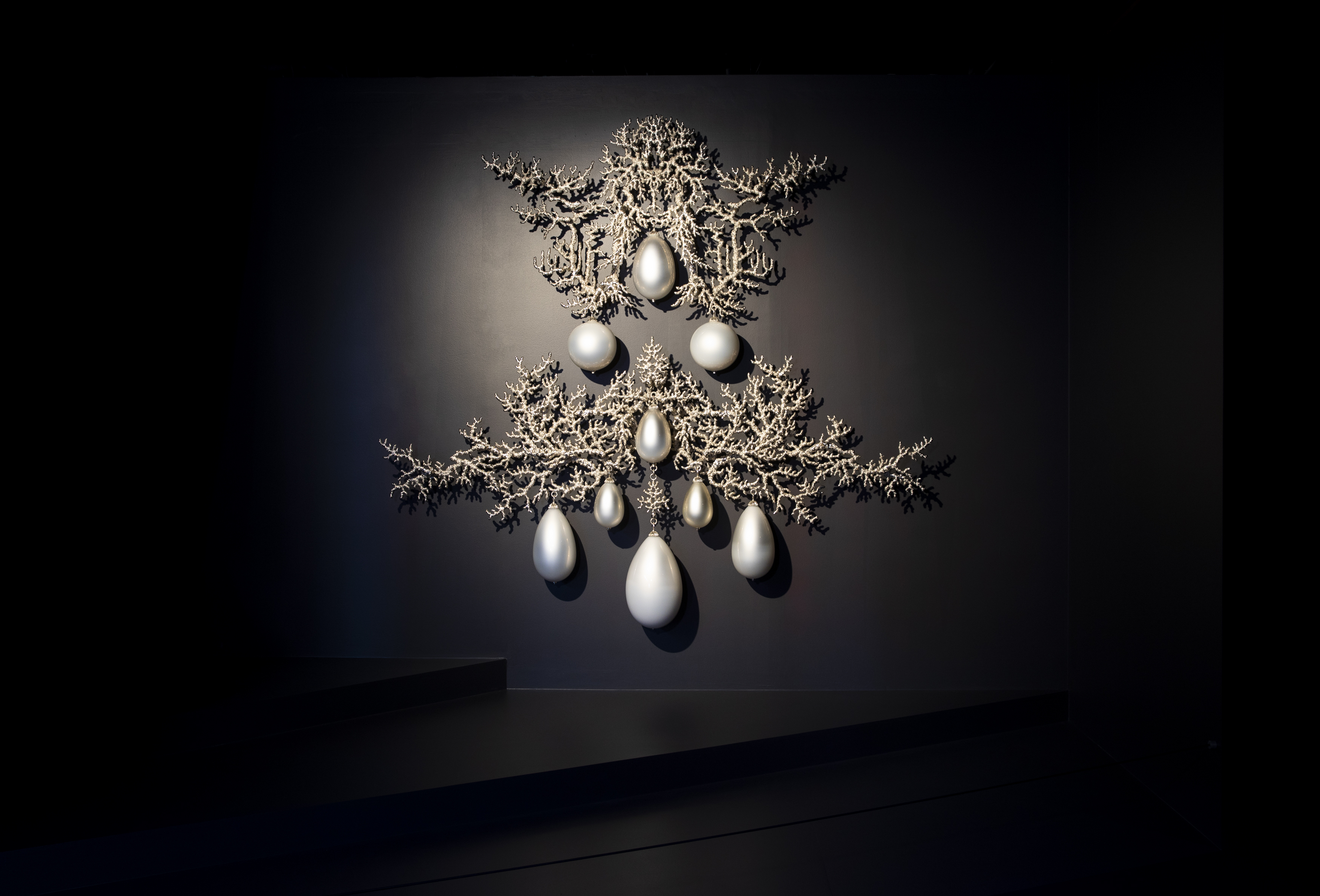 An ornate artwork in silver and white hangs on a black wall. It is in the form of a large symmetrically designed earring formed from silvery coral-like branches set with tear-shaped white glass pendants that look like pearls.